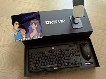 A black "OKX VIP" branded box and an anime-style trading card sit on a wooden desk, while below it, an opened black box displays a sleek keyboard and mouse set—showcasing the OKX VIP merchandise kit the user is excited to unbox. The text "OKX VIP" is clearly visible on the closed box, tying the premium trading platform's branding to the keyboard and mouse promotion.