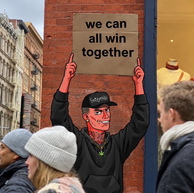 A street art piece on a brick wall depicting a man in a black cap and hoodie with his arms raised in a celebratory gesture, positioned below a cardboard sign reading 'we can all win together.'