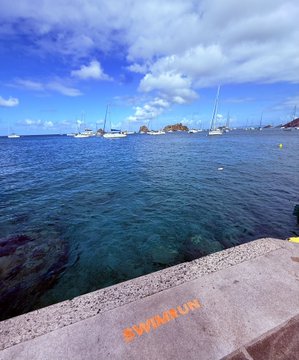 A scenic harbor view with sailboats moored in clear turquoise water under a partly cloudy sky, taken from a stone pier labeled 'SWIMMING' in orange paint.