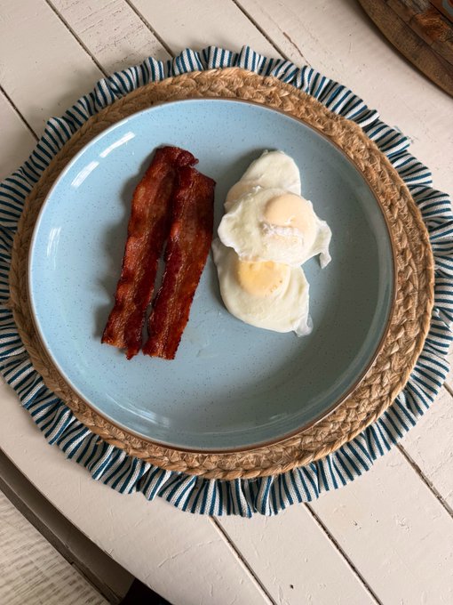A top-down view of a breakfast plate on a woven placemat, featuring two sunny-side-up eggs and two strips of crispy bacon on a light blue plate.