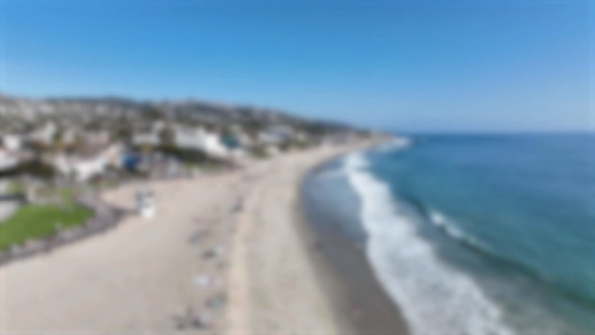 The image shows a beach scene with people enjoying their time near the water. The sky above them appears hazy, giving the impression of an overcast day. In the background, there are several houses along the shoreline, indicating that this is likely a residential area or a vacation spot for locals and tourists alike.