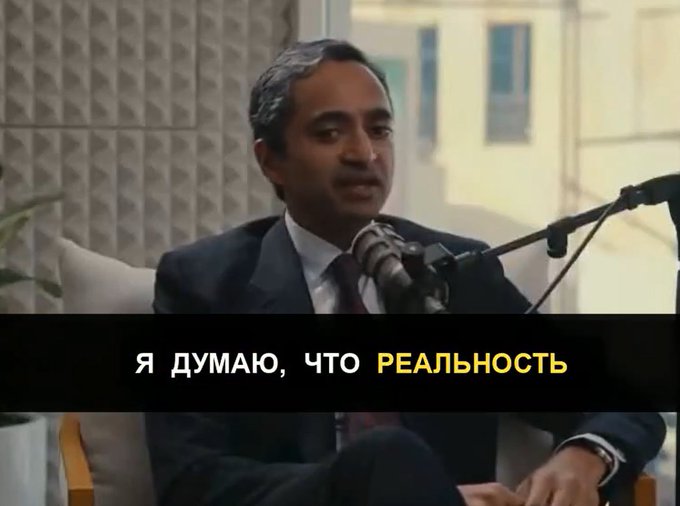 A man in a dark suit speaks into a microphone in what appears to be an interview setting, with Russian subtitles reading 'Я ДУМАЮ, ЧТО РЕАЛЬНОСТЬ' (I think that reality).