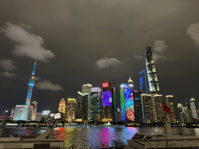 Nighttime cityscape of Shanghai's Pudong skyline across the Huangpu River, featuring illuminated skyscrapers including the Oriental Pearl Tower and Shanghai Tower, with colorful light reflections on the water.