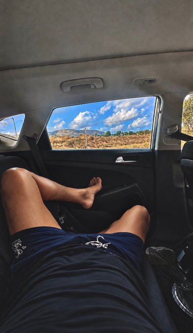 A person relaxes barefoot in the back seat of a car with their legs stretched out, wearing navy shorts, looking out the window at a scenic landscape of mountains and clouds.
