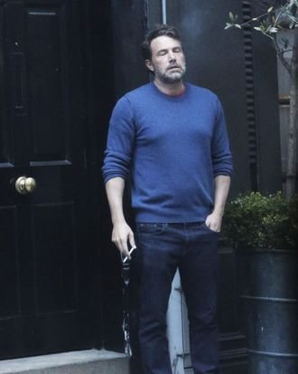 A candid photo of a man with a beard wearing a blue sweater and dark pants, standing by a black doorway holding what appears to be a leash.