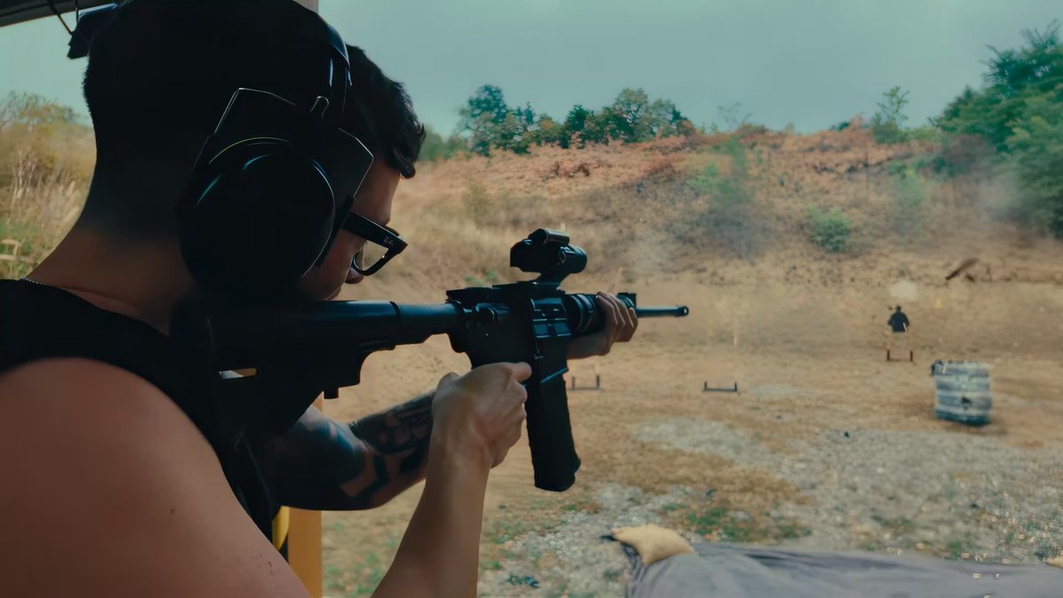 A person wearing ear protection and glasses aims a rifle at a target range with a dirt berm in the background.