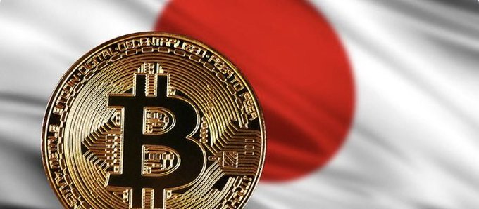 A close-up of a gold Bitcoin (₿) physical coin displayed in front of a blurred Japanese flag background.