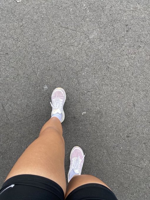 A first-person view of someone wearing lavender Nike sneakers and black shorts standing on cracked asphalt, perfectly capturing the "go outside touch some grass" weekend vibe.
