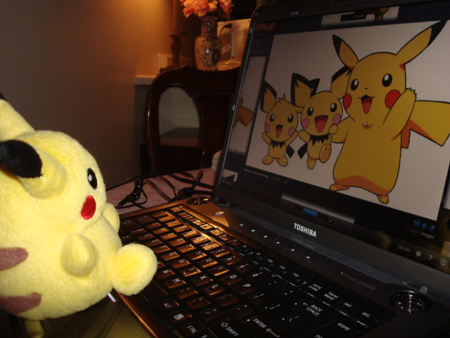 A yellow Pikachu plush toy sits next to a Toshiba laptop displaying an image of three Pikachu characters on its screen.