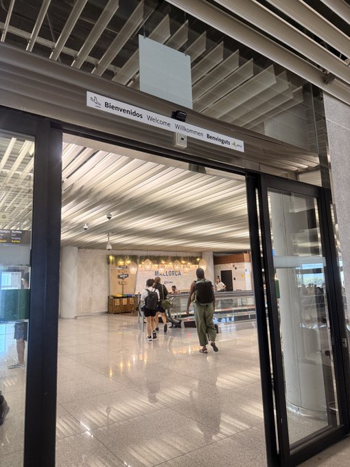 An airport terminal entrance displays a 'Bienvenidos Welcome Willkommen Benvinguts' sign above the doorway, with travelers walking toward a 'MALLORCA' information desk.