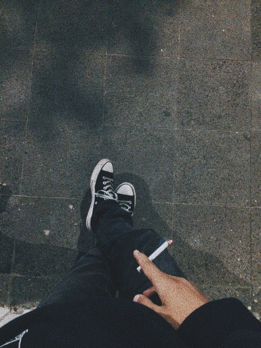 A moody nighttime photo taken from above showing a person's legs in black pants and black Converse sneakers on dark pavement, with their hand holding a cigarette visible in the frame.