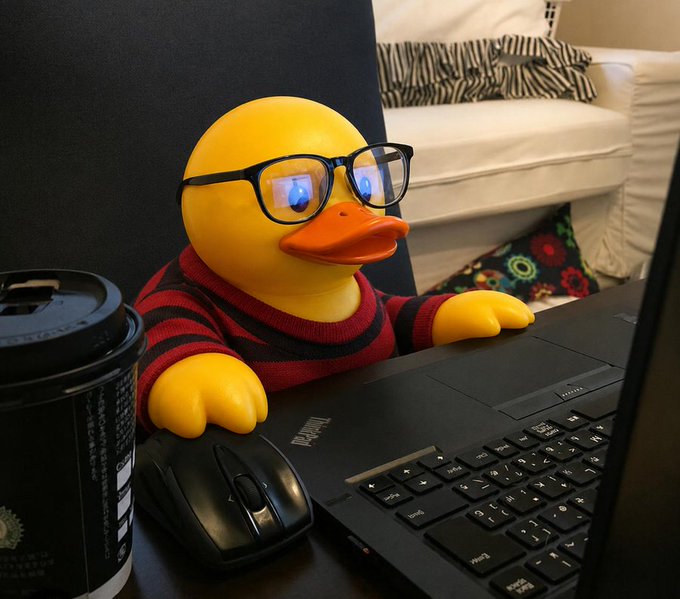 A rubber duck wearing black-framed glasses and a red-and-black striped sweater, positioned at a desk with a black laptop, a computer mouse, a coffee cup, and an office chair in the background.