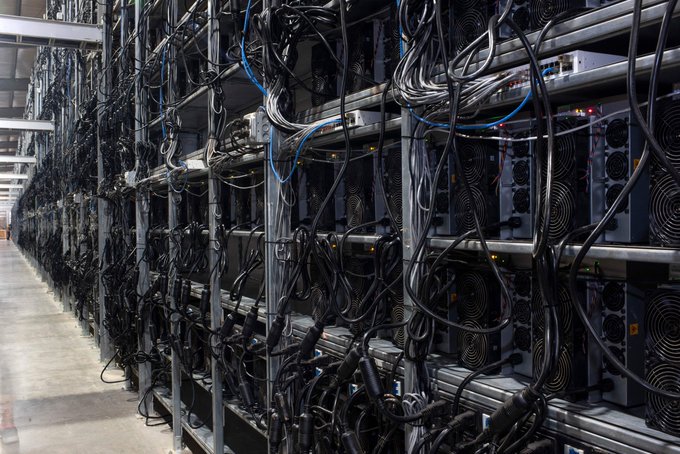 Rows of cryptocurrency mining hardware are densely packed on metal shelving units in a large warehouse, with numerous power cables connecting the machines.