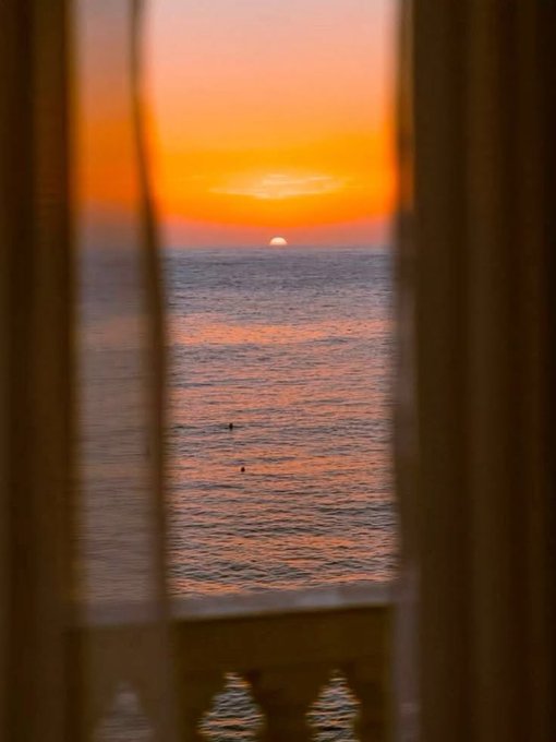 A breathtaking sunrise over the ocean, viewed through a window with curtains drawn aside, with the sun just cresting the horizon and casting a warm orange glow across the water—capturing the peaceful start to a "Happy Monday" morning.