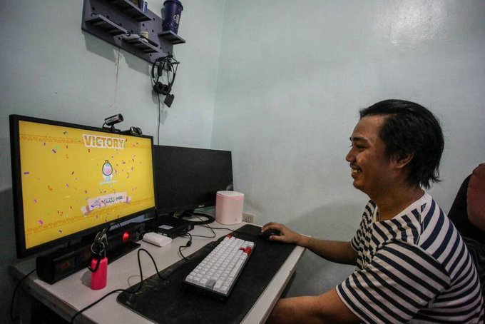 The image shows a man sitting at his desk, smiling while playing a video game on his computer. The man appears to be enjoying himself as he plays the game. There are two keyboards visible near him, one of which has a red keypad. A mouse can also be seen in front of the keyboard. In addition to these peripherals, there is a TV screen and a cup placed nearby.