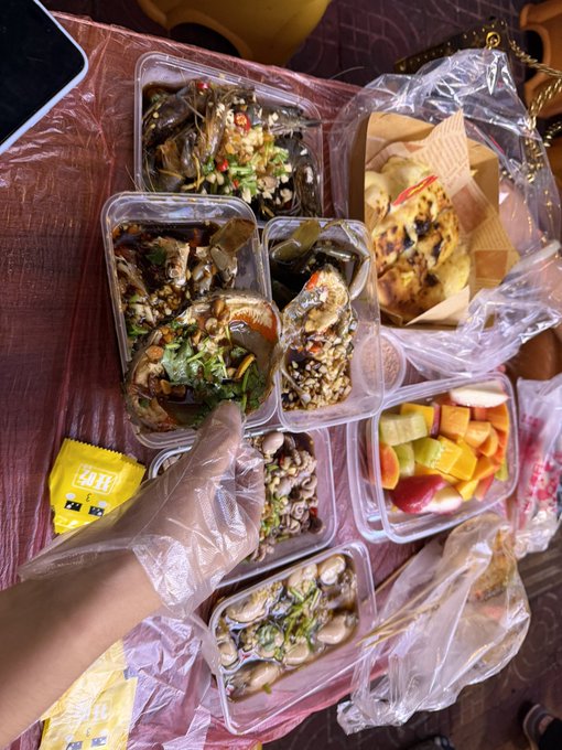 A hand in a plastic glove holds a takeaway spread of multiple plastic containers filled with marinated crab, shellfish, and chopped fruit, plus a paper bag of fried snacks. The post "潮汕啦～" (referring to Chaoshan, China) suggests this is a local specialty meal, likely featuring the region's famous marinated crab dishes.