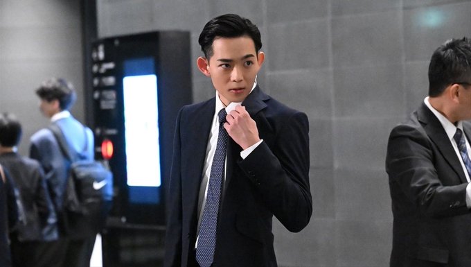 A man in a dark suit adjusts his patterned tie while standing in what appears to be a corporate lobby, with another suited man walking past on the right side of the frame.