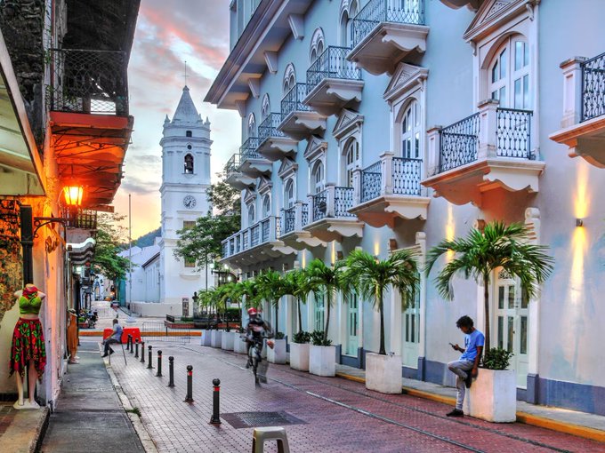 A charming colonial street in Casco Viejo, Panama, featuring a clock tower, pastel blue buildings with wrought-iron balconies, palm trees, and locals walking through at sunset, complementing the post's message about Panama as an emerging haven for Bitcoiners seeking freedom and alternative residency.