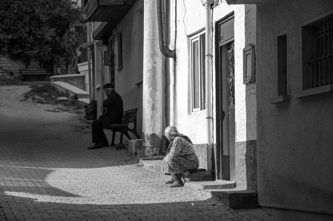 Black and white street photograph of two men sitting along a building exterior on a cobblestone street, with one crouched near the steps and another seated on a bench.