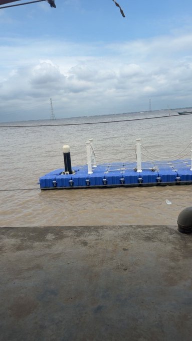 A blue floating pontoon dock with white posts and a black-topped mooring bollard sits in muddy brown water under a cloudy sky, with electrical transmission towers visible on the distant shoreline.