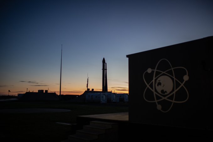 A silhouette of a rocket on a launchpad at sunset, next to a building featuring an atom logo containing a globe.