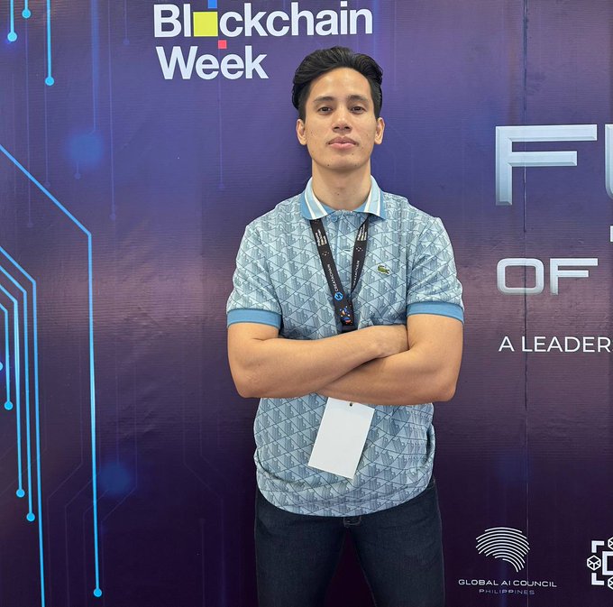 A young man in a patterned polo shirt stands with crossed arms at a 'Blockchain Week' event, wearing a lanyard with an ID badge, against a backdrop with 'FUTURE OF...' and 'GLOBAL AI COUNCIL PHILIPPINES' logos.