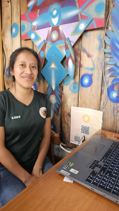 A smiling woman wearing a black 'LUNY' shirt sits at a wooden desk with a Lenovo Legion laptop, with a QR code display and a colorful painted wooden wall behind her.