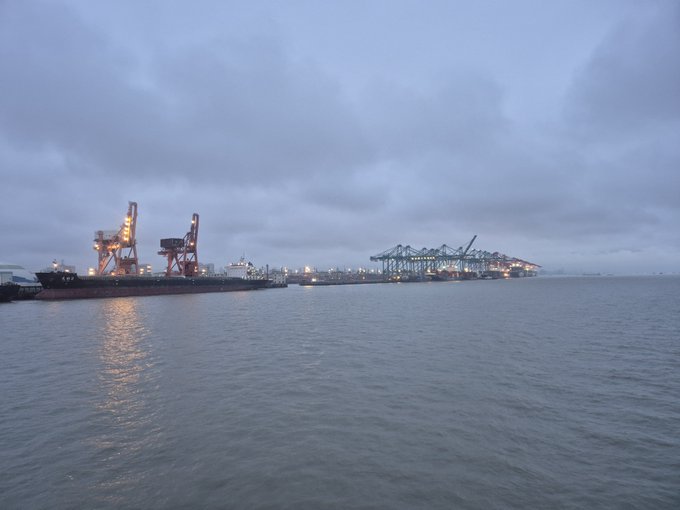 An early morning or evening view of a port with cranes, shipping containers, and cargo ships across the water under a cloudy sky.