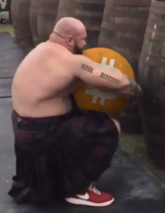 A meme image showing a shirtless, bald, heavily built man kneeling and holding a large orange coin featuring the Bitcoin logo, with a 'HODL BITCOIN' tattoo on his arm.