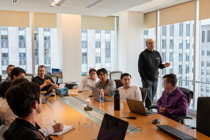 A presenter addresses a focused group of professionals around a conference table, surrounded by laptops and engaged in what appears to be a high-stakes trading or business meeting. The image visually reinforces the post's sentiment by depicting the collaborative, attentive team environment behind the impressive individual performances highlighted in the message.