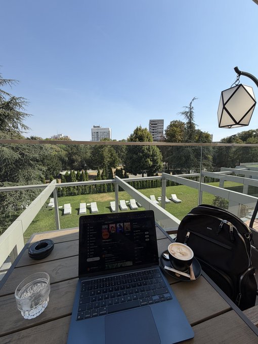A scenic outdoor workspace on a wooden deck overlooking a garden and city skyline, featuring an open laptop, a glass of water, a coffee cup, headphones, and a laptop bag.
