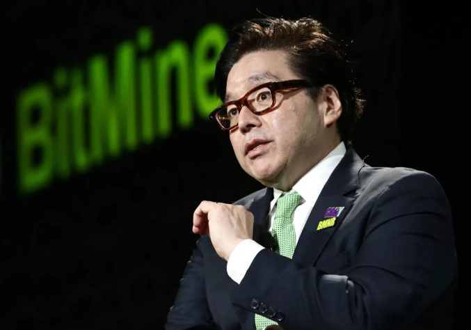 A man in a dark suit, green tie, and glasses is shown mid-speech with the "BitMine" logo displayed in green behind him. The image relates to the post by highlighting Tom Lee's leadership of Bitmine Immersion Technologies as the company continues its large-scale Ethereum accumulation strategy, aiming to secure 5% of the global ETH supply.