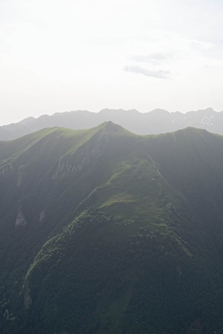 A misty, hazy aerial view of green mountain ridges and forested peaks under a pale, overcast sky creates a serene landscape scene.