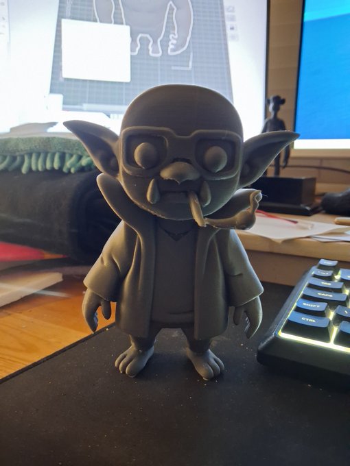 A gray 3D-printed figure of a goblin-like character wearing glasses and a robe, sitting on a desk next to a glowing RGB keyboard.