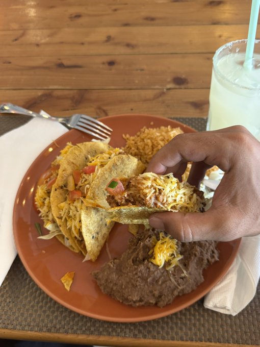 A hand picks up a cheese-topped hard taco from a plate piled with Mexican food, including rice, refried beans, and additional tacos, accompanied by a fork and a glass of lemonade on a wooden table—visually embodying the "Taco Tuesday" celebration referenced in the post about the $.13 $Sol purchase and rug pull question.