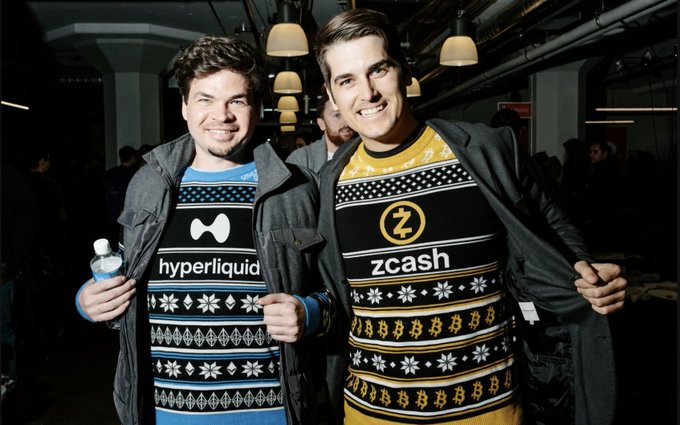 Two men posing together wearing Christmas-style sweaters, one with a 'hyperliquid' logo and the other with a 'zcash' logo featuring the Z symbol.