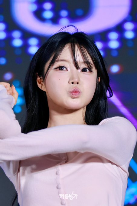 A young woman with dark hair in a light pink long-sleeved top poses with a playful pouting expression, arms raised, against a stage background with blue lights. Korean text is visible at the bottom.
