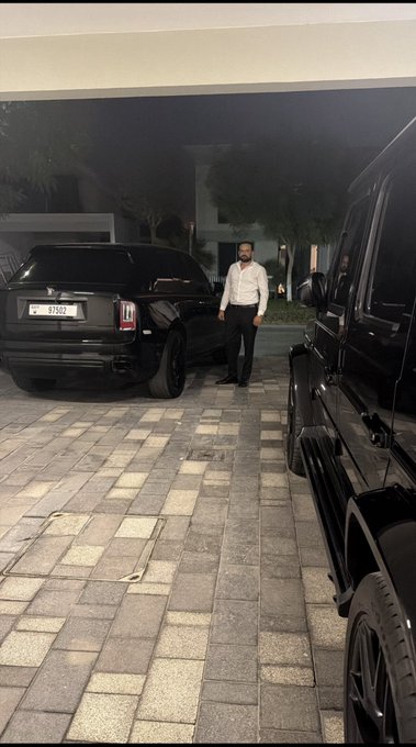 A nighttime scene of a man in a white shirt and black pants walking between two black SUVs, with one car displaying license plate '83902'.