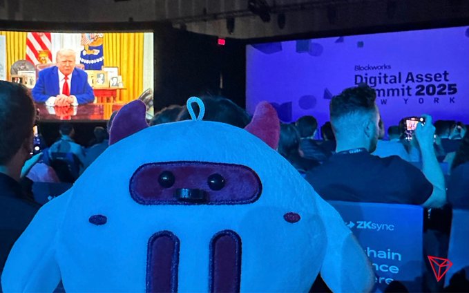 At a 'Blockworks Digital Asset Summit 2025' event, a person in a furry mascot costume sits in the audience watching a broadcast of Donald Trump speaking.