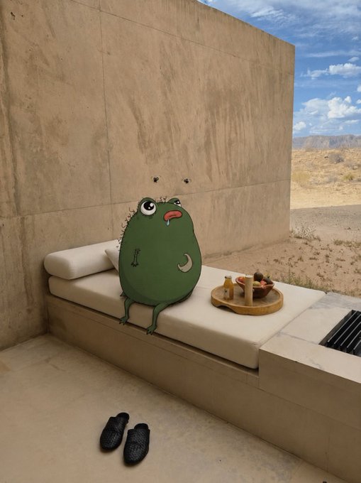 A green cartoon monster sits on a beige cushioned bench against a concrete wall in a desert landscape, with a tray of fruit and drinks nearby and a pair of woven shoes on the ground.