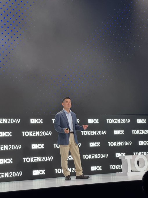 A man in a blue blazer and khaki pants giving a presentation on stage at TOKEN2049, standing in front of a backdrop featuring the event's logo repeated across it with blue dotted lights above.