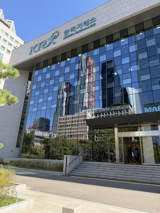 The image shows the exterior of the Korea Exchange (KRX) building, with its logo and name "한국거래증권거래소 KOREA EXCHANGE" prominently displayed above the reflective glass facade. The building's mirror-like windows capture reflections of surrounding skyscrapers, marking the venue of the introduction event for the Korea Exchange.