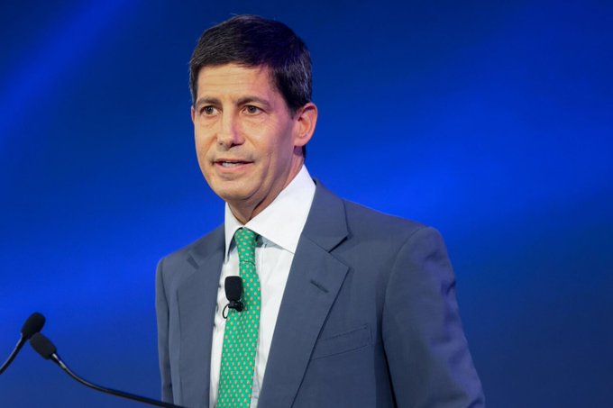 A man in a dark suit and green tie speaks at a podium with a microphone against a blue background.
