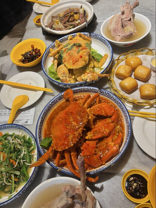 A top-down view of a large Chinese banquet table featuring a prominent whole crab in a spicy orange sauce, accompanied by fried soft-shell crabs, steamed buns, and various other dishes.