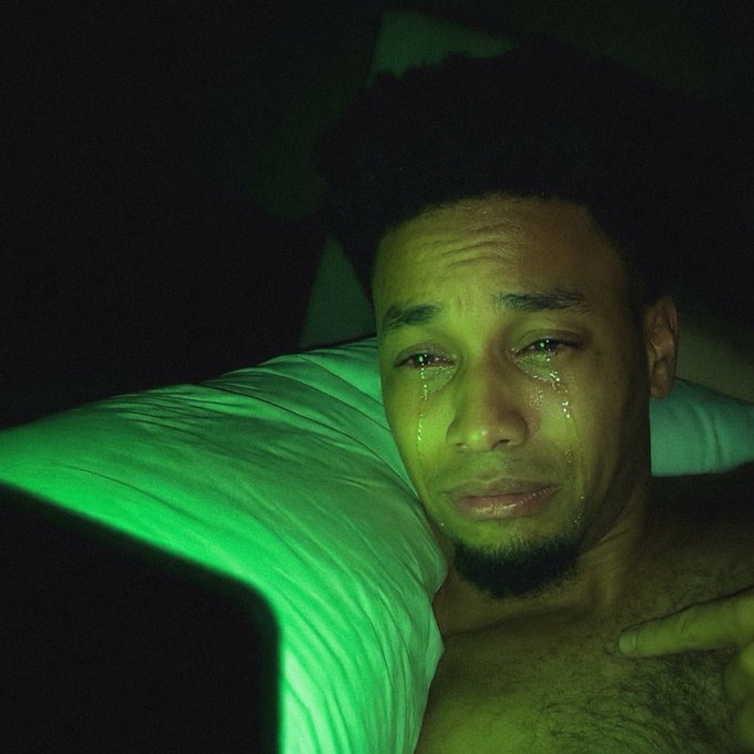A man lies in bed with tears streaming down his face, illuminated by a green glow.