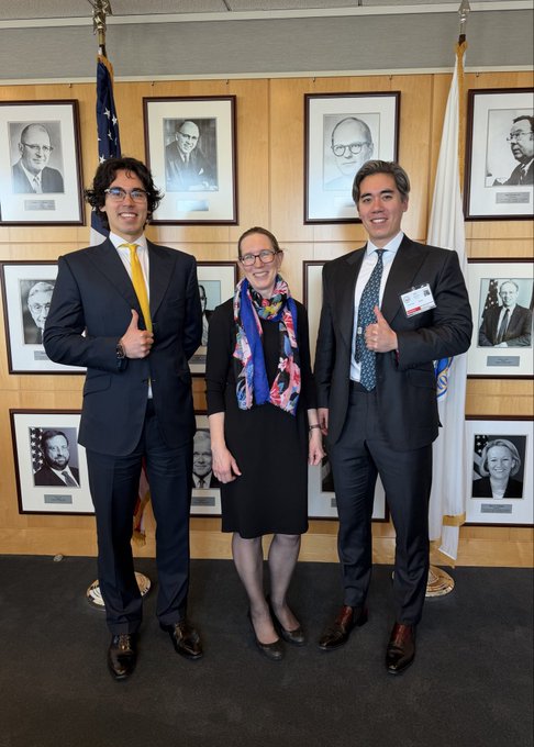 Three professionally dressed individuals pose for a photo in front of a wall featuring framed portraits of notable figures, with one giving a thumbs-up gesture.