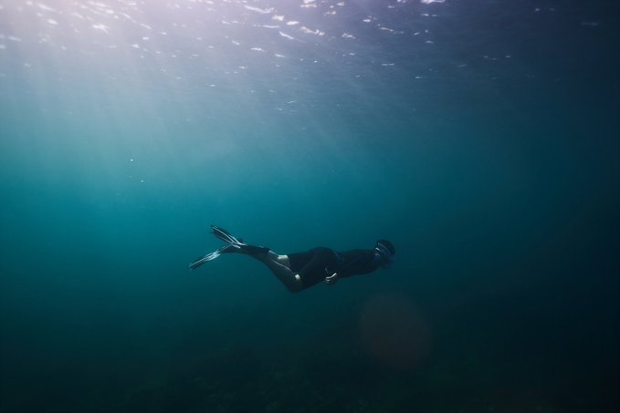 A person in a wetsuit with fins snorkels underwater in deep teal water, with sunlight streaming down from the surface above.