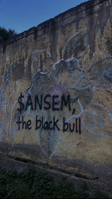 The image shows a weathered, moss-streaked concrete wall with blue spray-painted graffiti reading "$ANSEM, the black bull." This matches the post, which describes the user painting "$ANSEM" on their grandfather's old wall.