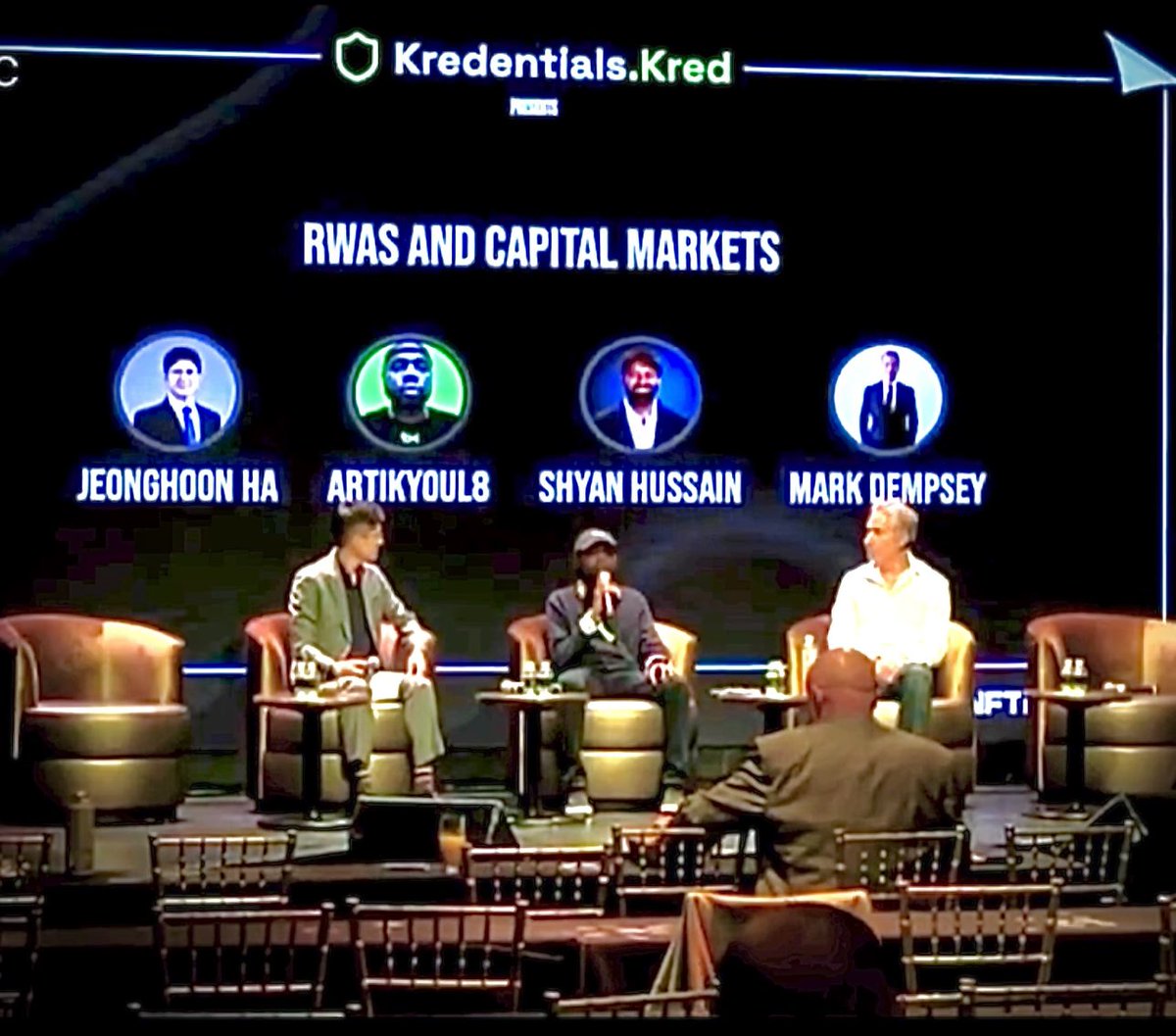 A photograph of a panel discussion titled 'RWAS AND CAPITAL MARKETS' hosted by Kredentials.Kred, showing three people seated on a stage below circular portraits labeled 'JEONGHOON HA', 'ARTIKYOUL8', 'SHYAN HUSSAIN', and 'MARK DEMPSEY'.
