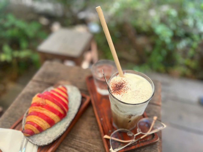 An outdoor cafe scene featuring an iced coffee with a bamboo straw and cocoa powder dusting, served next to a colorful red and orange croissant on a wooden tray.